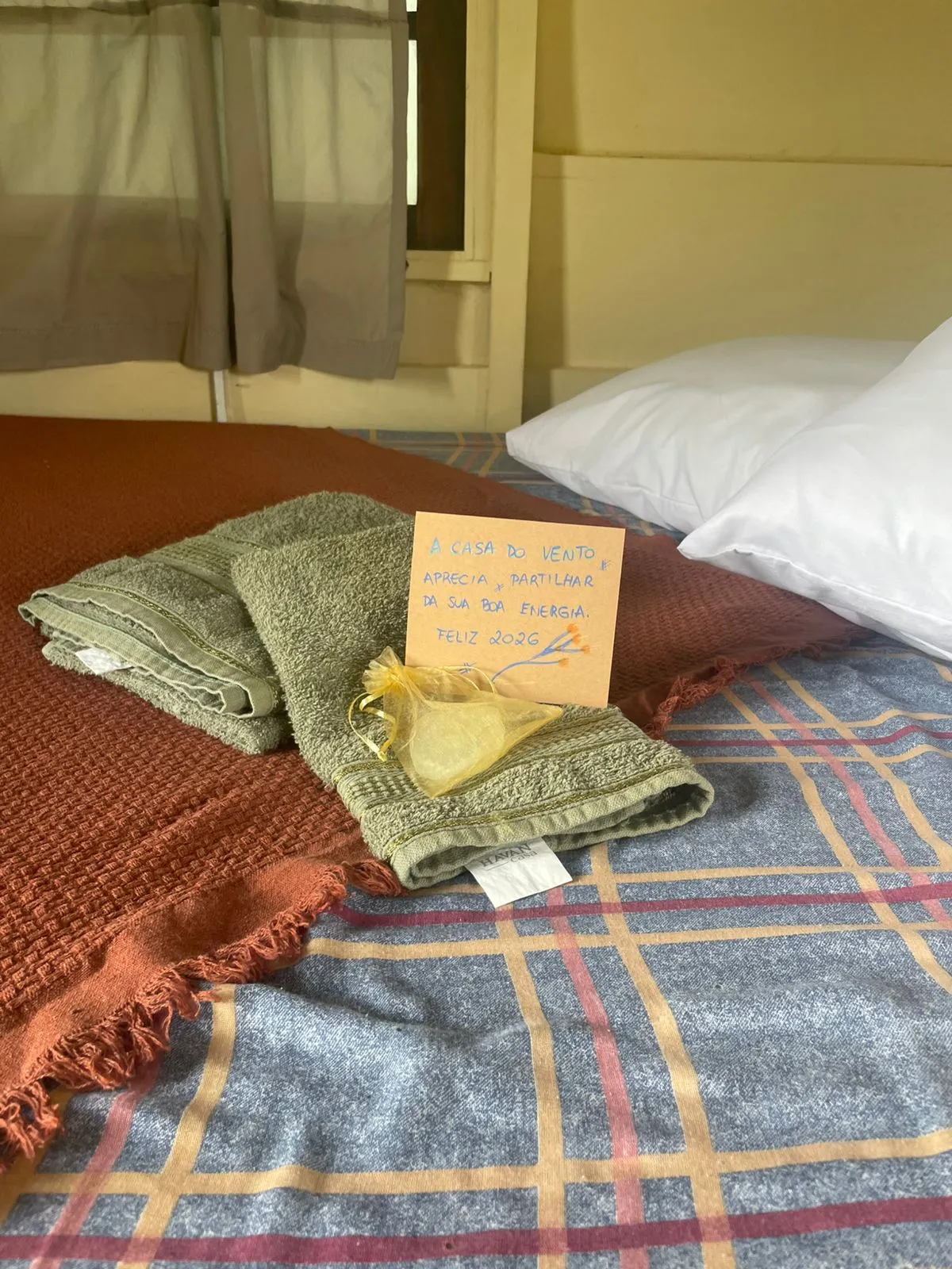 Cama de hospedeiro com toalhas dobradas, sabonete artesanal e bilhete de boas-vindas com votos de boas energias