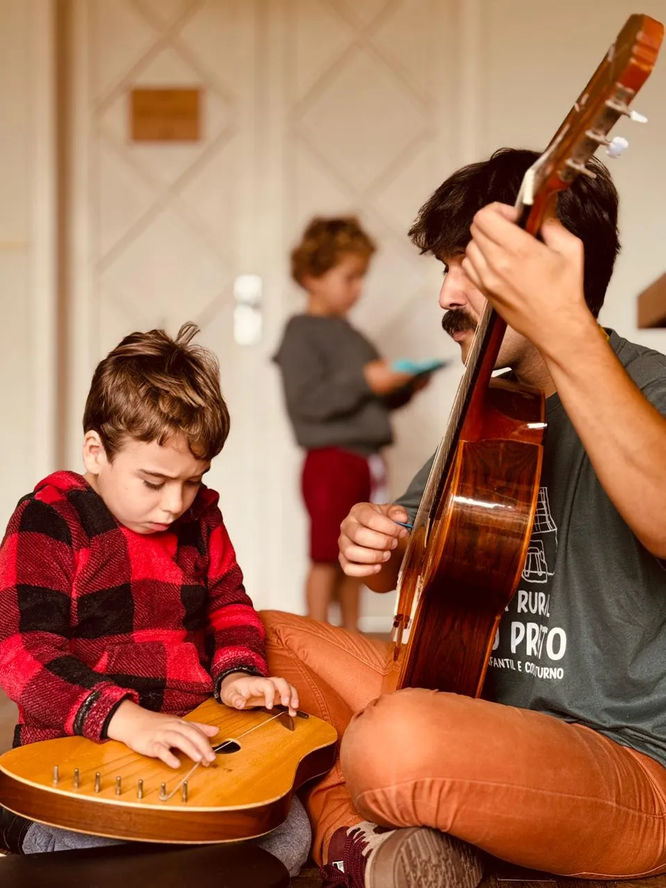 Menino concentrado tocando um instrumento de cordas deitado nas pernas de um músico com violão